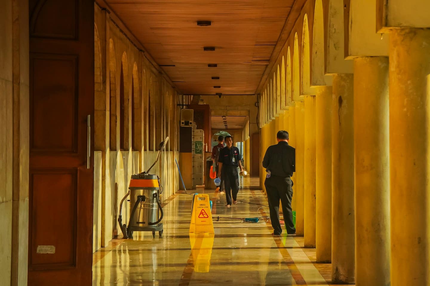 Maintenance workers clean a long hallway with a wet floor sign.